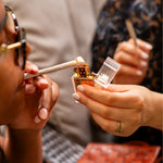 A close-up lifestyle shot of a person lighting a cigarette with a gold lighter held in their hand. The lighter has a polished gold body, a clear plastic cap, and a visible flame.
