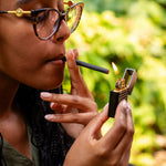 A close-up lifestyle shot of a person lighting a black clove cigarette with a black lighter held in their hand.