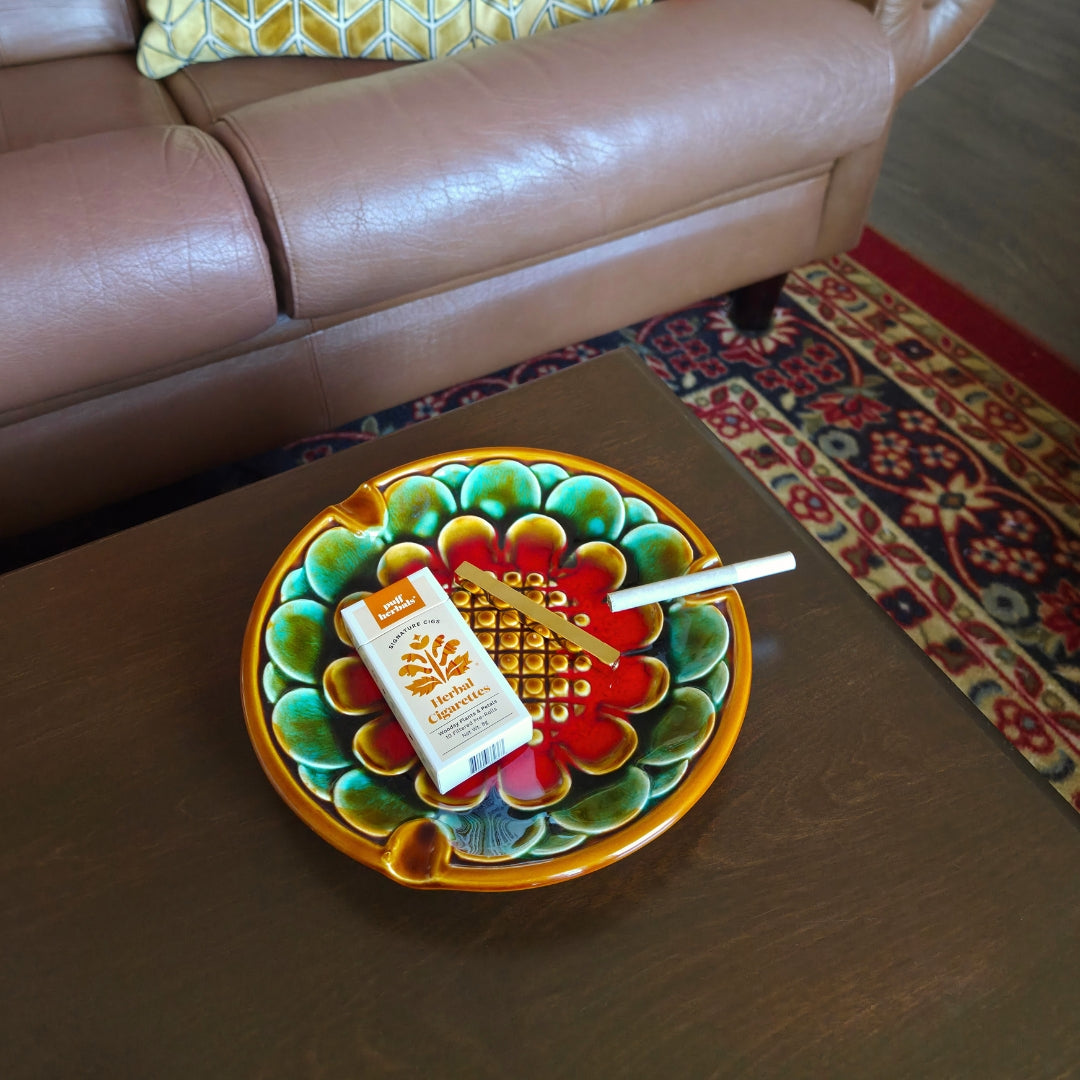 Colorful decorative plate with herbal cigarettes on a wooden table, with a brown sofa and patterned rug in the background.