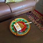 Colorful decorative plate with herbal cigarettes on a wooden table, with a brown sofa and patterned rug in the background.