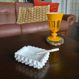 White textured ashtray with a cigarette on a wooden table, yellow lamp in the background
