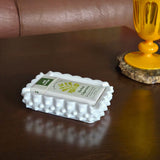 White textured box with a label on a wooden surface next to a yellow candle holder.